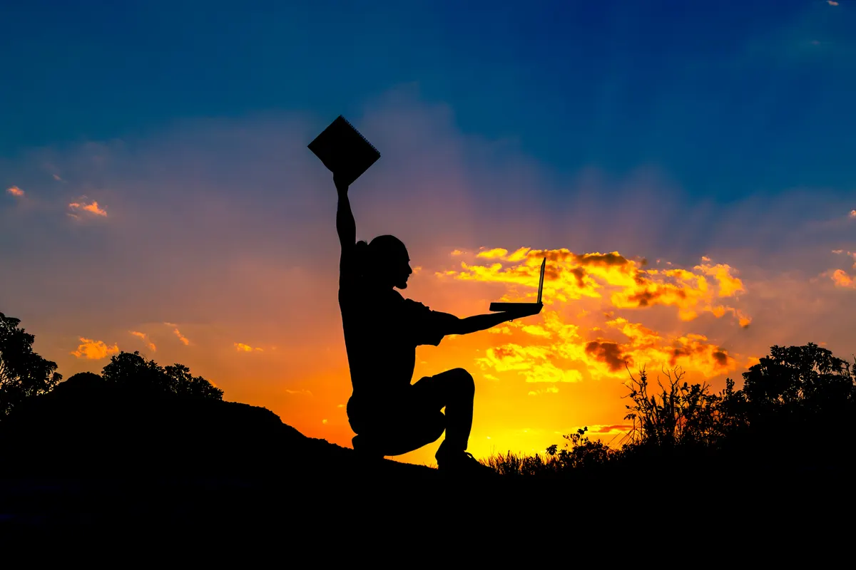 Silhueta de Arthur ao pôr do sol, erguendo um caderno numa mão e segurando um notebook na outra