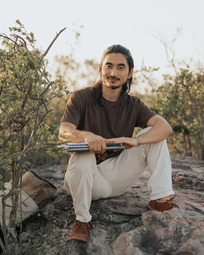 Arthur Kubagawa sentado sobre uma pedra, ao ar livre, sorrindo